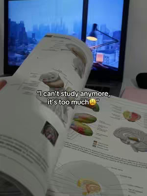 The image shows a person holding open a textbook with colorful illustrations. The image is taken from a low angle, looking up at the book and the person's hands. Behind the book, there's a laptop showing a blurred-out city scene. Overlaid on the open book is text, a quote expressing study fatigue.