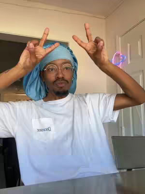 A person with dark skin is positioned in front of a blurry indoor background. The person is wearing a pale blue bonnet, round glasses, and a white t-shirt. The person's hands are raised, displaying a peace sign. The person has short facial hair.