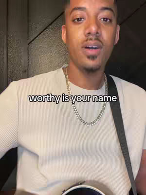 A medium close-up shot of a man singing with his mouth open and looking up slightly. He's wearing a light-colored, ribbed shirt, a silver chain, and a black guitar strap is visible over his shoulder. The text overlay 'worthy is your name' is displayed at the bottom of the frame. The background is a dark wooden panel.