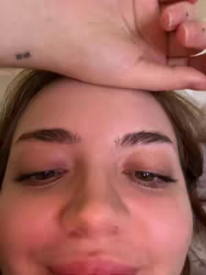 A close-up shot features a person's face. The main focus is on the eyes, which are closed or nearly closed. Their eyebrows are well-defined and arched, adding to the expression. A wrist with a small tattoo is visible, and a hand is placed over the head. The skin tone appears light, and the overall lighting suggests an indoor setting.