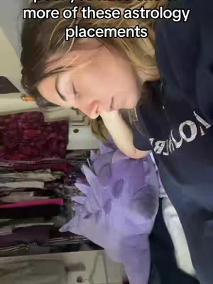 A woman appears to be lying down with her head resting on a purple plush object, possibly a pillow or stuffed toy. The background reveals a cluttered space with folded clothes in the back, and the woman is wearing a black top with some text partially visible. Text overlayed at the top reads 'more of these astrology placements.'