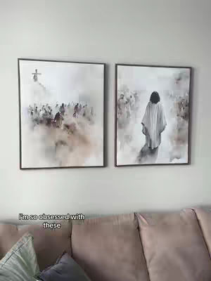 The image shows two framed artworks hanging on a wall, depicting artistic interpretations of religious scenes. Below, part of a plush sofa is visible, suggesting an interior setting.