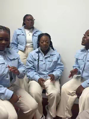 A group of five women in light blue jackets and white pants are seated or standing against a plain white background. The central figures are singing, with facial expressions and hand gestures emphasizing the performance.
