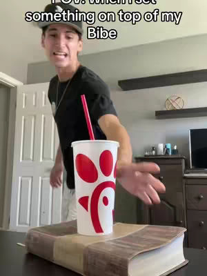 A young man in a black t-shirt stands in a room with a Chick-fil-A cup and a red straw placed on top of a Bible. He gestures towards the cup with his right hand as if showing it off. The background shows a light-colored wall. Underneath him is a bible placed on a table.