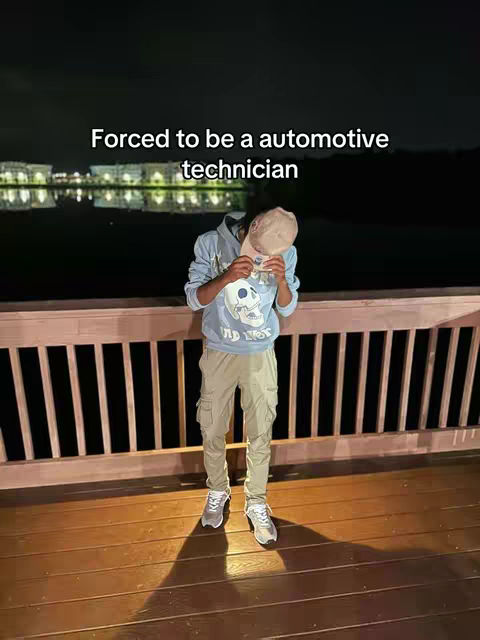 A person wearing a light blue hoodie, khaki pants, and sneakers stands on a wooden deck, with their face obscured by a cap. The background features a blurred night scene with lights reflecting on water and a wooden railing in the mid-ground. The caption above the head says: Forced to be a automotive technician.