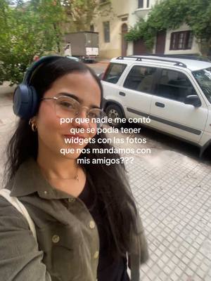 A selfie-style shot with a young woman as the central figure. She wears glasses and blue headphones, smiling slightly. The background features a street scene with a white car parked on the right and buildings with greenery, suggesting a daytime setting. Text overlay on the image, in Spanish, focuses on widgets with photos with friends.