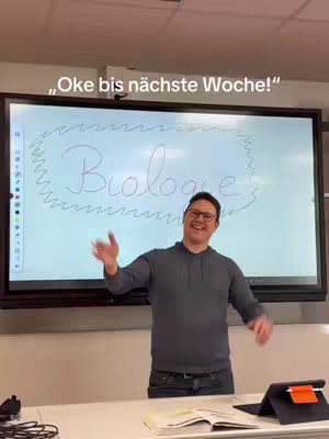 A man in glasses stands and gestures with open arms in front of a whiteboard that reads 'Biologie' in a handwritten style, within a wavy border.