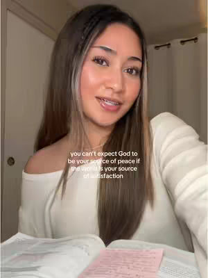 A woman with long brown hair and a white off-the-shoulder top is looking at the camera and smiling. Overlayed in white text: "you can't expect God to be your source of peace if the world is your source of satisfaction". She is sitting in what appears to be a room with a neutral-colored wall and a door in the background and a page with pink post-it notes lies open on the table in front of her.