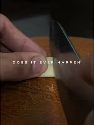 A close-up shot of a person's hand using a knife to cut a piece of garlic on a wooden surface. Overlayed text asks 'DOES IT EVER HAPPEN'. The lighting is somewhat dim, suggesting an indoor setting.