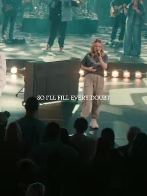 A live performance shot with a female singer center stage, holding a microphone and singing. The stage is lit with warm lighting, and there is a band and audience visible in the background.