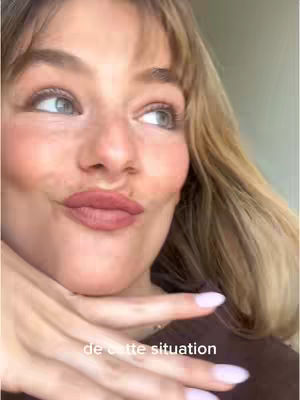 The cover image shows a close-up of a woman's face. She has fair skin, blonde hair with bangs, and is looking to the side with her lips slightly pursed. Her hand is in front of her mouth suggesting she is deep in thought or reacting to something. Her nails are painted in a light pink color. The background is a blurred neutral tone.