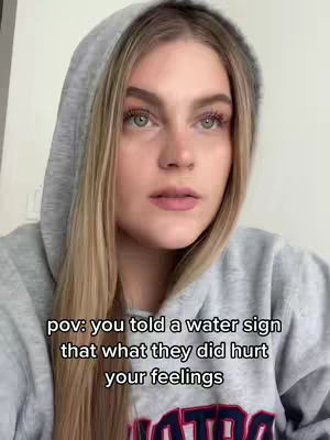 A young woman with blonde hair and a gray hoodie is looking directly at the camera with a thoughtful expression. Text overlays describe a scenario related to a water sign's feelings.