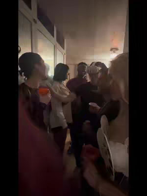 A group of people are gathered inside with a casual atmosphere; most individuals are holding drinks or small plates; the scene appears to be a social gathering or party, showcasing potential demonstrations of augmented reality.