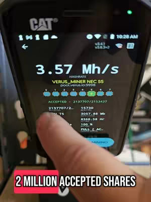 A close-up shot of a black CAT-branded smartphone displaying a mining interface. A finger is pointing at the screen near the bottom where a red banner displays the text: '2 MILLION ACCEPTED SHARES'. The screen shows real-time data, including the hash rate, network information, and the number of accepted shares. The phone is slightly angled, and the background is out of focus.
