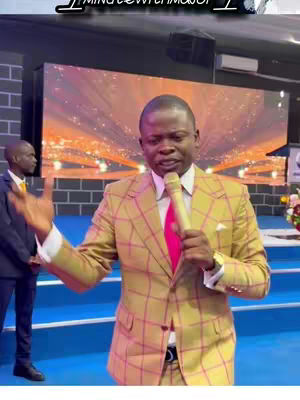 A man in a gold checkered suit holds a microphone, speaking in what appears to be a church setting. A second man stand in the background with a black suit and a yellow tie. Behind them there is a led screen where the image is a blurry sun-like display.