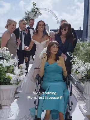 A group of people, including a bride in a wedding dress, are walking down a paved path with white flowers on either side, a woman in a wheelchair in a blue dress is being pushed. The background is blurred, suggesting they are outdoors on a sunny day.