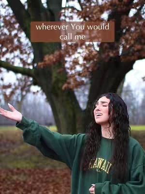 A woman with long dark curly hair stands outdoors with her arms outstretched, head tilted back in what appears to be a moment of praise or singing. She wears a green sweater, and the scene is set against a blurred backdrop of trees with reddish-brown leaves and a muted sky. A brown rectangle with white text is superimposed in the upper part of the image.