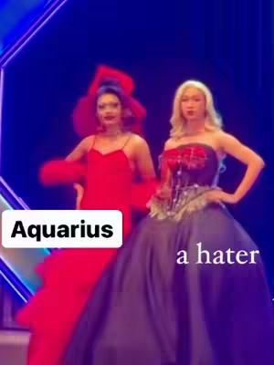 Two individuals in elaborate gowns stand on a stage with a dark blue background. The person on the left wears a long red dress topped with a red headpiece and flowing sleeves. The person on the right is in a strapless gown, with a dark bodice and a purple skirt. Text overlays read 'Aquarius' in a white box and 'a hater' in the lower right.