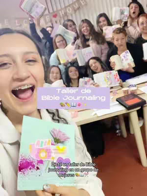 A selfie style shot featuring a woman in the foreground, smiling widely with visible braces. She is holding a decorated bible journaling page, with several people holding bibles and journaling materials in the background with high energy.