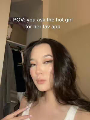 A selfie-style video shows a young woman, framed from the chest up, in a dimly lit room. She has long, dark hair and is looking towards the camera with a slight smile. She is dressed in a white top, and her expression suggests she is responding to a question. The background includes a partially visible open closet space and neutral-toned walls, with the focus on the subject. The text overlay reads "POV: you ask the hot girl for her fav app".