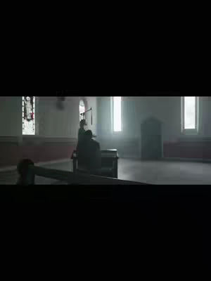 The image captures a dimly lit interior, possibly a church. Light streams through tall windows, illuminating a central space where figures are present. The composition is simple, with a focus on the architecture and the people or objects within the room.