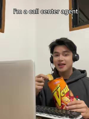 A young man wearing a headset and a dark gray hoodie is holding an open bag of chips in one hand and a chip in the other, smiling at the camera. A laptop and keyboard create a home office setting and the text overlay reads: 'I'm a call center agent.'