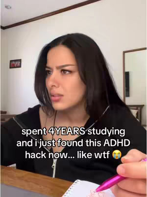 A woman with long dark hair looks at the camera with a concerned expression. Text overlayed on the image reads, "spent 4 YEARS studying and I just found this ADHD hack now... like wtf 😭". She appears to be holding a pen and writing in a notebook. The background includes a wall, corner, and what appears to be a mirror.