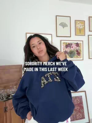 A woman in a dark blue hoodie with an 'AXO' design stands in front of a wall with framed pictures. The text overlay reads 'SORORITY MERCH WE'VE MADE IN THIS LAST WEEK'. The background includes a wooden cabinet and decor.