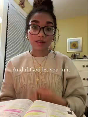 A young woman with glasses and her hair up is speaking while holding an open book. The background is a room with a window with blinds and a picture on the wall.