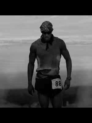 A monochrome image of a man running outdoors, possibly on a mountain or hill. He is wearing a bandana, sunglasses, a tank top, and shorts with a number pinned to them. The background is blurred, probably sky.