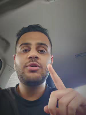 A man with short dark hair and a trimmed beard is pointing his right index finger up towards the right. He is seated in what appears to be a vehicle, as the background is the car ceiling. The man is wearing a black shirt.