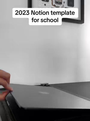 The cover image shows a close-up shot of a black desk with a laptop, and a hand is seen partially visible on the left side. Above the workspace, there's a framed piece of art on a white wall. Text overlaid on the image reads '2023 Notion template for school'.