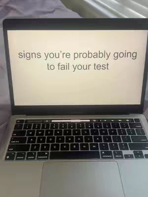 A Macbook is open, with the screen displaying the text: "signs you're probably going to fail your test". The keyboard and the bottom half of the screen are visible. The background is blurred.