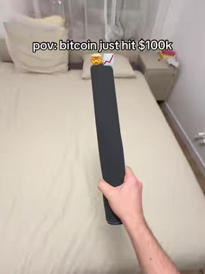 A first-person perspective inside a bedroom. A hand holding a black object, possibly a phone or a smart device, is pointing towards the ceiling. The bed is covered with a light-cream comforter, and two pillows are seen. The overall lighting shows a bright ambience. The words 'pov: bitcoin just hit $100k' is seen as well as an emoji of money bags, and a stock chart.
