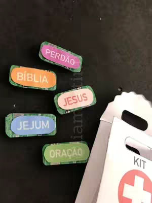The image shows a collection of religious-themed craft items against a dark surface. Several small, colorful rectangular labels with rounded edges are scattered across the scene. Each label features a different word in bold, white text against a brightly colored background: 'PERDAO' (forgiveness) in pink, 'BIBLIA' (Bible) in orange, 'JESUS' in light pink, 'JEJUM' (fasting) in blue, and 'ORACAO' (prayer) in green. These elements appear to be part of a 'kit' or organized set, as a white, handle-bag is visible. The bag features the word 'KIT' and a red cross symbol, suggesting a first-aid or spiritual kit.