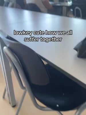 The image shows a classroom setting with chairs and a table. The text overlay says, "lowkey cute how we all suffer together". A chair against a table is in the foreground, while other chairs are visible in the background.