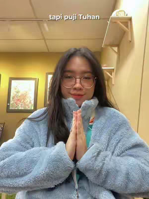 A selfie featuring a young woman with long black hair wearing a light blue fleece jacket, glasses, and a neutral expression, with her hands clasped in front of her chest. The background shows a warmly lit interior with framed pictures on the wall.