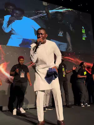 A man in a white suit sings into a microphone during an event. The background features a projected image of the performer and others. The image is well-lit, emphasizing the central figure dressed in a white outfit.