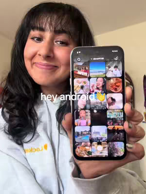A young woman with dark hair and bangs is smiling while holding a smartphone displaying a grid of photos. She's wearing a light gray hoodie. The background is a blurred interior.