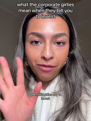 A selfie-style video image featuring a light-skinned woman with long wavy hair, wearing a white sweater and gold hoop earrings. She is looking directly at the camera and holding up her hand with her palm forward. Overlayed text in the upper left reads, 'what the corporate girlies mean when they tell you to invest,' and text at the bottom of the frame reads, 'when the girlies say to invest.' The background is blurred.