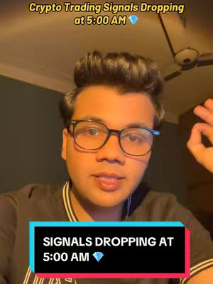 A young man with glasses is in the center of the frame, looking towards the camera with a slightly concerned expression. Above the man is a text that says "Crypto Trading Signals Dropping at 5:00 AM" in yellow font. Below that, there's a rectangular text overlay in black that states "SIGNALS DROPPING AT 5:00 AM" with a diamond emoji and a bright blue background. The lighting suggests an indoor setting, and the composition focuses on the man's upper body and face.