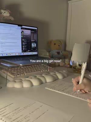 A well-lit desk setup at night with a laptop open displaying a video-streaming app, a pen in hand ready to write on a paper, along with stationary and a few stuffed toys on the desk.