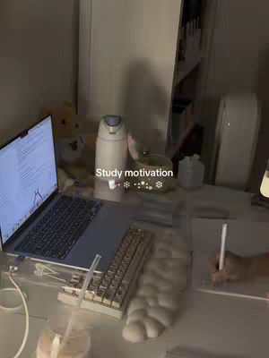 A dimly lit, slightly cluttered desk setup. A laptop sits open on the left, displaying text. A white keyboard and what appears to be a drawing tablet are in front of it. To the right, a hand is holding a pen over a sheet of paper. In the background, a teddy bear, a water bottle, and other desk supplies are visible. The overlay text 'Study motivation' is in the center of the image.