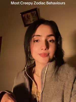 A young woman with dark hair and fair skin is the focal point. She's wearing a gray top and has headphones in. The background is a softly lit room with a small framed picture above her head.