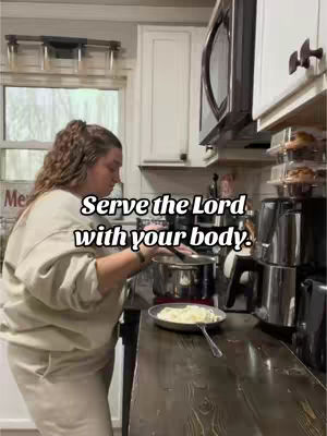 A woman is in a kitchen, seen from the side while cooking at the stove. The setting includes white cabinets and a window that peers into the outside. The overlay text reads, 'Serve the Lord with your body.'