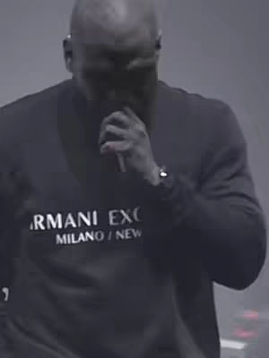 The cover image features a medium shot of a dark-skinned man with a shaved head, dressed in a black long-sleeved shirt. He's holding a microphone close to his mouth, and the overall lighting suggests a dimly lit environment. The image has a somewhat grainy quality.