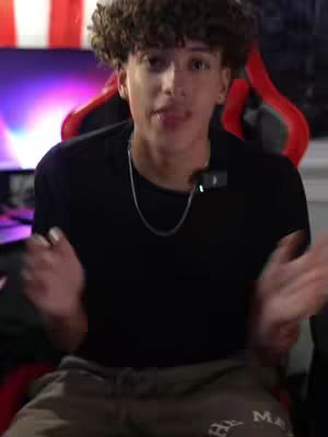 A young man with curly hair sits in a red and black gaming chair with arms outstretched. He wears a black t-shirt and silver chain. The background is slightly blurred, but the colors purple and red dominate. A small microphone is clipped to his shirt.