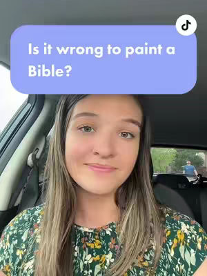 A young woman with long, light brown hair is seated inside a car. She wears a green and yellow floral top and smiles. Above her head, a light blue rectangle contains the text: "Is it wrong to paint a Bible?" In the background, a blurry scene suggests an outdoor setting, possibly a residential area.