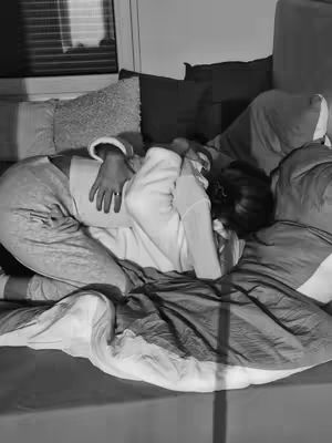 A black and white photo shows a couple embracing in bed, creating a cozy and intimate scene. The composition features a bed with rumpled sheets and pillows, and the lighting appears soft and natural. Overall, the image conveys a sense of comfort, love, and closeness between the couple.