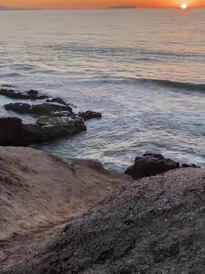 A scenic view of a coastline at sunset. The image is dominated by the vast expanse of the ocean, meeting the sky at the horizon in a gradient of colors from orange to yellow. The water has gentle waves. In the foreground on the bottom, there is a rocky cliff face, textured in shades of brown and gray.